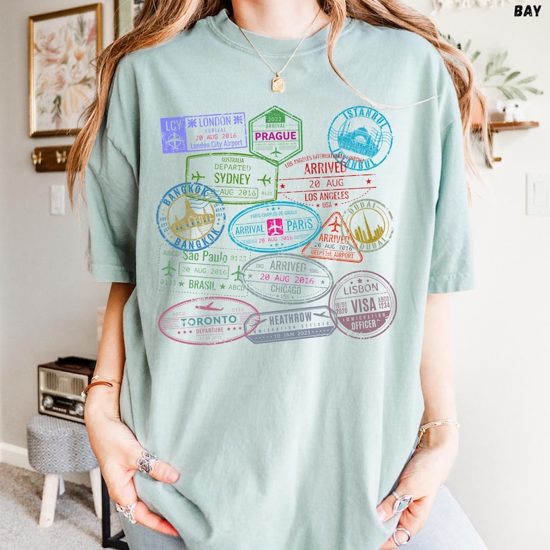 以下が含まれることがあります： 薄い青色のTシャツに、ロンドン、プラハ、イスタンブール、シドニー、バンコク、パリ、ドバイ、サンパウロ、シカゴ、リスボン、トロントなど、世界中のさまざまな都市のカラフルなパスポートスタンプのグラフィックプリントが施されています。スタンプには、到着日と出発日、都市名、空港コードが記載されています。