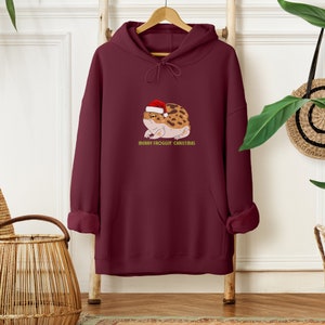 Op de afbeelding: Bordeauxkleurige hoodie met een zak en een afbeelding van een kikker met een kerstmanmuts. De tekst "Merry Frogg'in Christmas" staat onder de kikker.