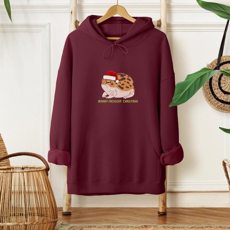 Op de afbeelding: Bordeauxkleurige hoodie met een zak en een afbeelding van een kikker met een kerstmanmuts. De tekst "Merry Frogg'in Christmas" staat onder de kikker.