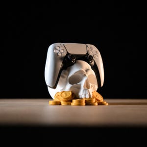 May include: A white human skull sculpture resting on a pile of gold-colored circular coins, with a white and black video game controller balanced on top of the skull. The entire object is set against a dark, solid black background.