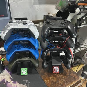 Pu&ograve; includere: Due pile di controller per videogiochi. Una pila presenta un controller mimetico bianco e grigio, controller blu e verde oliva. L'altra pila ha controller neri. Ogni pila poggia su una base di ricarica con un logo della console.