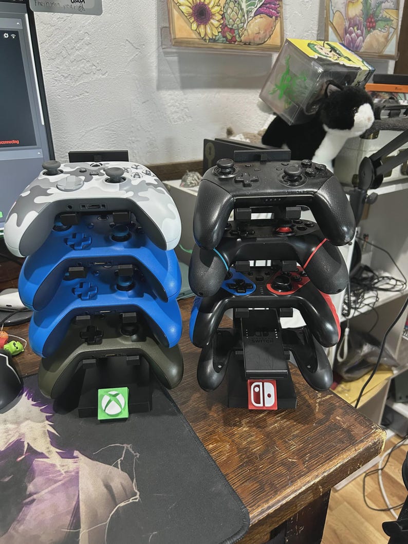 Pu&ograve; includere: Due pile di controller per videogiochi. Una pila presenta un controller mimetico bianco e grigio, controller blu e verde oliva. L'altra pila ha controller neri. Ogni pila poggia su una base di ricarica con un logo della console.