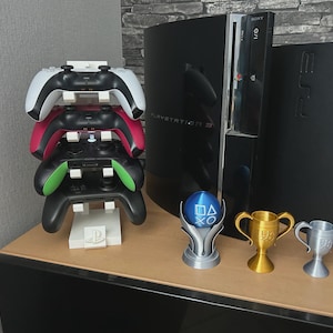 Pu&ograve; includere: Una pila di quattro controller di gioco bianchi, rossi, verdi e neri, appoggiati su un supporto bianco con un logo PlayStation. Una console PlayStation 3 e diversi trofei sono sullo sfondo.