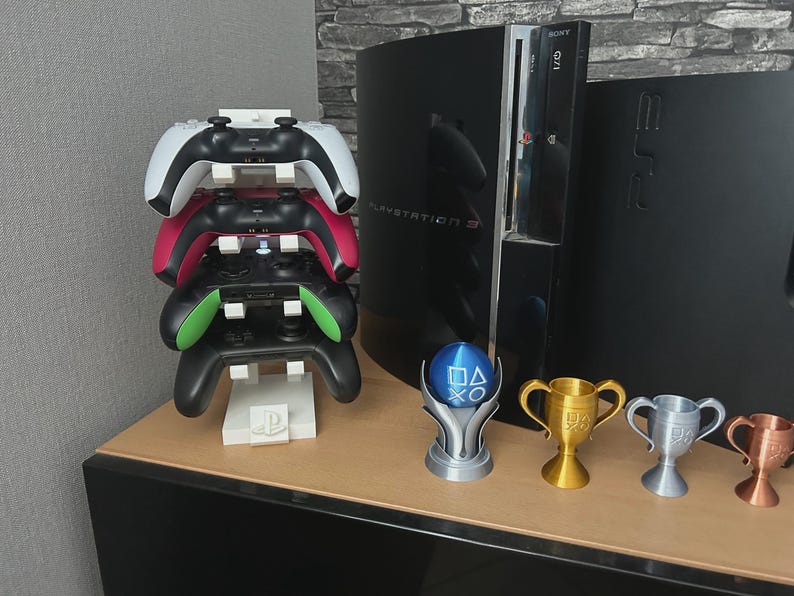 Pu&ograve; includere: Una pila di quattro controller di gioco bianchi, rossi, verdi e neri, appoggiati su un supporto bianco con un logo PlayStation. Una console PlayStation 3 e diversi trofei sono sullo sfondo.