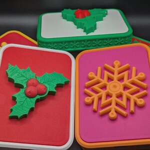 May include: A collection of Christmas-themed boxes with festive designs. The boxes feature a red, white, and pink base with green holly, red berries, and orange snowflakes. The boxes have a white border and are set against a dark background.
