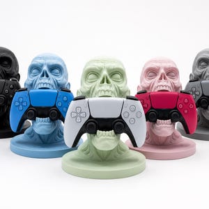 May include: A collection of five skull-shaped gaming controller stands in various colors: black, blue, green, pink, and gray. Each stand holds a matching colored controller, creating a striking visual display. The stands are designed to hold the controllers upright.