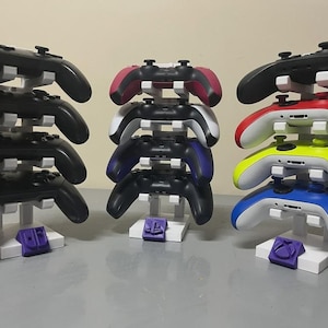 May include: Three white stands displaying multiple game controllers. The controllers are black, red, pink, blue, yellow, and white. Each stand has a coloured logo at the base.
