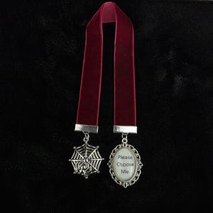 May include: A deep red velvet ribbon bookmark featuring two silver-toned metal charms. One charm is a spider web design, and the other is an oval pendant with a decorative filigree frame containing the text Please Choose Me. in black font.