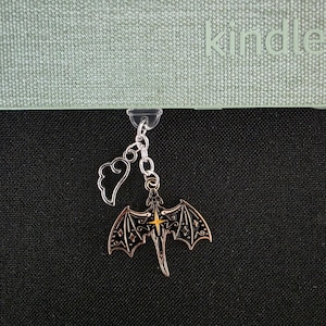 May include: A metal charm featuring a black and copper-colored dragon with an orange star detail, attached to a small silver-toned wing charm. The charm hangs from a clear plastic clip secured to the edge of a sage green Kindle e-reader case.