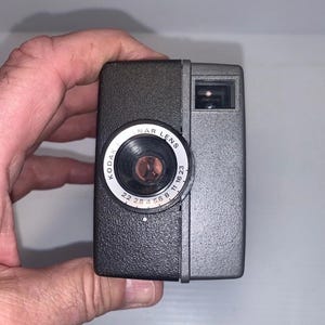 May include: A vintage Kodak camera with a dark gray body and a black lens. The lens is labeled "NAR LENS" with aperture settings. The camera is held in a person's hand.