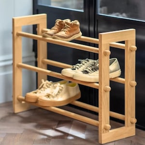 Puede incluir: Un zapatero de madera de tres niveles con tres pares de zapatos. El zapatero es de madera clara y tiene un diseño sencillo y minimalista. Los zapatos son de varios estilos y colores, incluyendo zapatillas deportivas y botas.