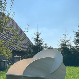 May include: A modern, sculptural wooden chair with a unique, curved design. The chair is constructed from light-colored wood and features a series of intersecting circular and semi-circular elements. It is set outdoors on a grassy lawn under a clear blue sky.
