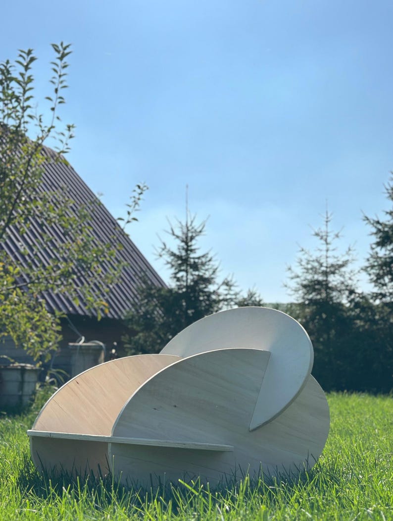 May include: A modern, sculptural wooden chair with a unique, curved design. The chair is constructed from light-colored wood and features a series of intersecting circular and semi-circular elements. It is set outdoors on a grassy lawn under a clear blue sky.