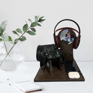 Puede incluir: Un soporte de juego de madera marrón oscuro con un soporte para auriculares incorporado, dos mandos de juego negros, un teléfono inteligente y un par de auriculares inalámbricos blancos. El soporte para auriculares tiene un recorte circular con una foto de una persona con auriculares. A la izquierda hay un jarrón de cristal transparente con una planta verde.