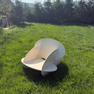 May include: A wooden chair with a unique, modern design. The chair is made of light-colored wood and features curved sides and a rounded back. It is placed on a grassy lawn with trees in the background.