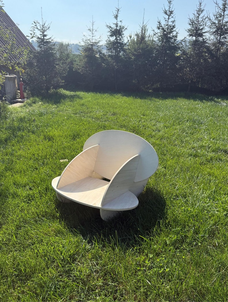 May include: A wooden chair with a unique, modern design. The chair is made of light-colored wood and features curved sides and a rounded back. It is placed on a grassy lawn with trees in the background.