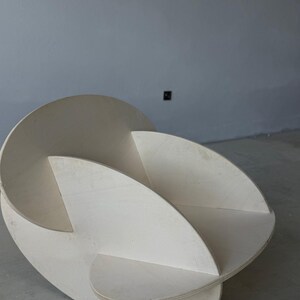 May include: A modern, abstract wooden sculpture with a light, natural finish. The piece features curved, intersecting planes, creating an open, three-dimensional form. The sculpture is designed to be a decorative art piece.