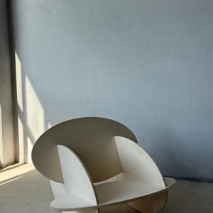 May include: A modern, sculptural chair constructed from light-colored plywood. The chair features a circular backrest and curved side supports, with a rounded seat. The design is minimalist and geometric, suitable for contemporary interiors.