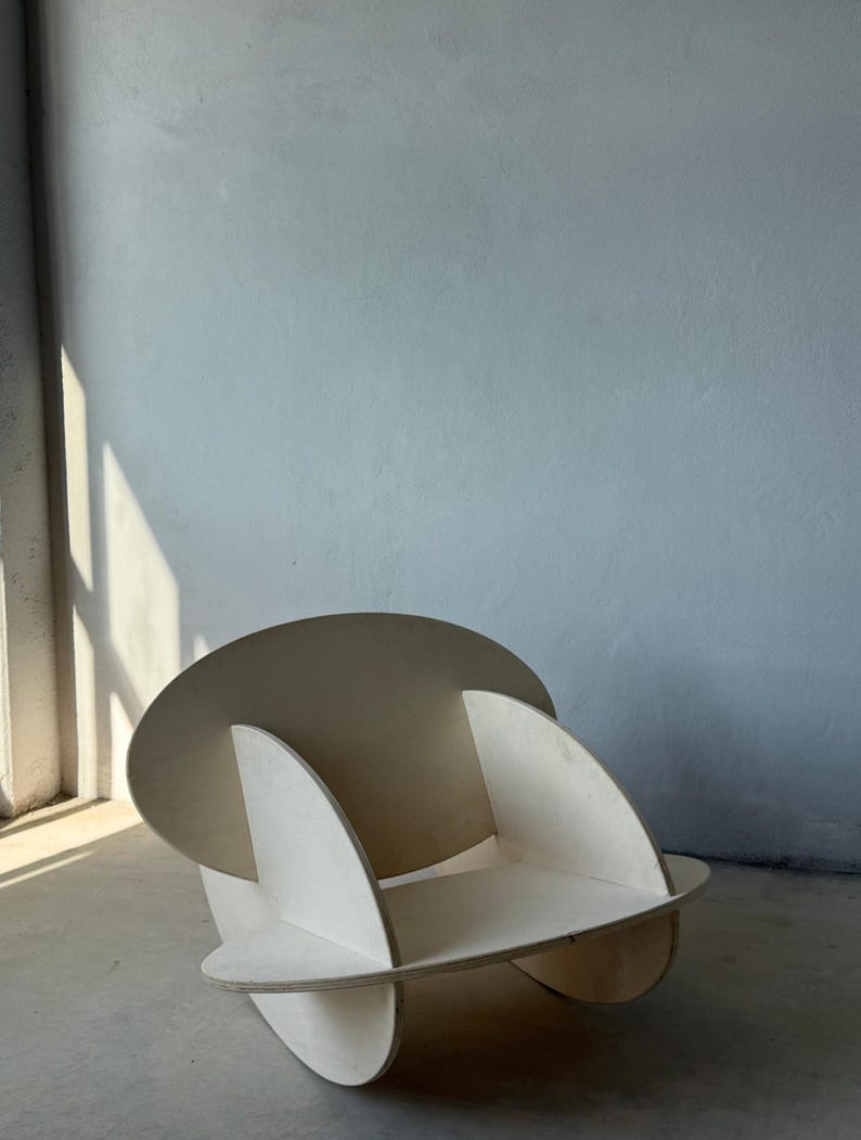 May include: A modern, sculptural chair constructed from light-colored plywood. The chair features a circular backrest and curved side supports, with a rounded seat. The design is minimalist and geometric, suitable for contemporary interiors.