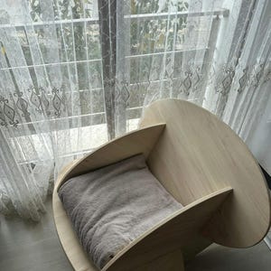 May include: A light-colored wooden pet bed with a curved design. The bed features a soft, gray cushion inside. The bed is placed near a window with sheer curtains.