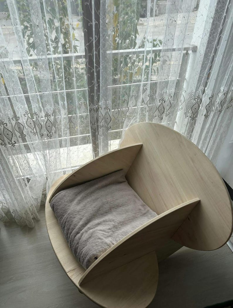 May include: A light-colored wooden pet bed with a curved design. The bed features a soft, gray cushion inside. The bed is placed near a window with sheer curtains.