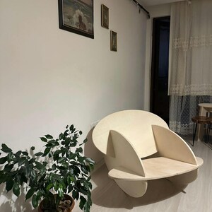 May include: A modern, light-colored wooden chair with a unique, rounded design. The chair has a circular back and curved armrests, with a flat seat. The chair is placed in a room with a plant and artwork on the wall.
