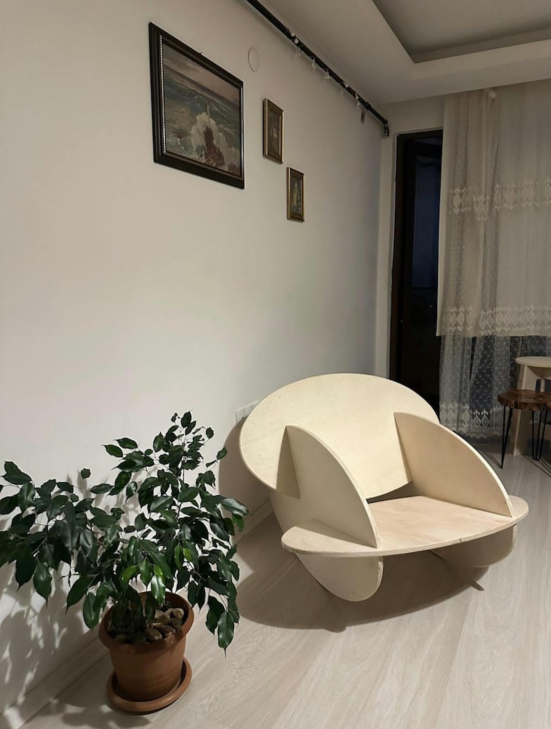 May include: A modern, light-colored wooden chair with a unique, rounded design. The chair has a circular back and curved armrests, with a flat seat. The chair is placed in a room with a plant and artwork on the wall.