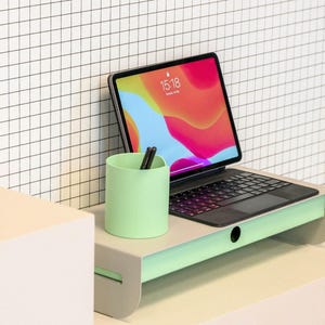 May include: A mint green desk organizer with a built-in shelf holds a tablet with a black keyboard case. A matching mint green pencil holder with black pens sits on the shelf. The background is a white grid pattern.
