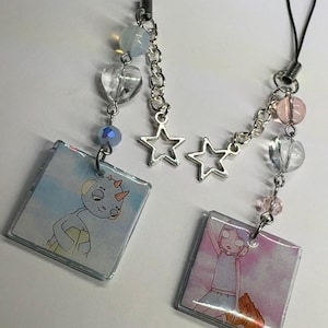 May include: Two square charms with cartoon characters on a silver chain with star and crystal beads. The charms are attached to a phone strap.