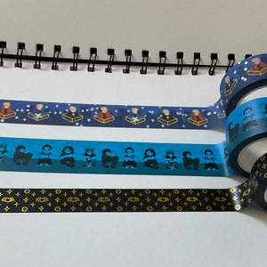 May include: Three rolls of blue and black patterned washi tape. The top roll has a pattern of small figures in boxes. The middle roll has a pattern of small figures. The bottom roll has a pattern of black and gold geometric shapes.