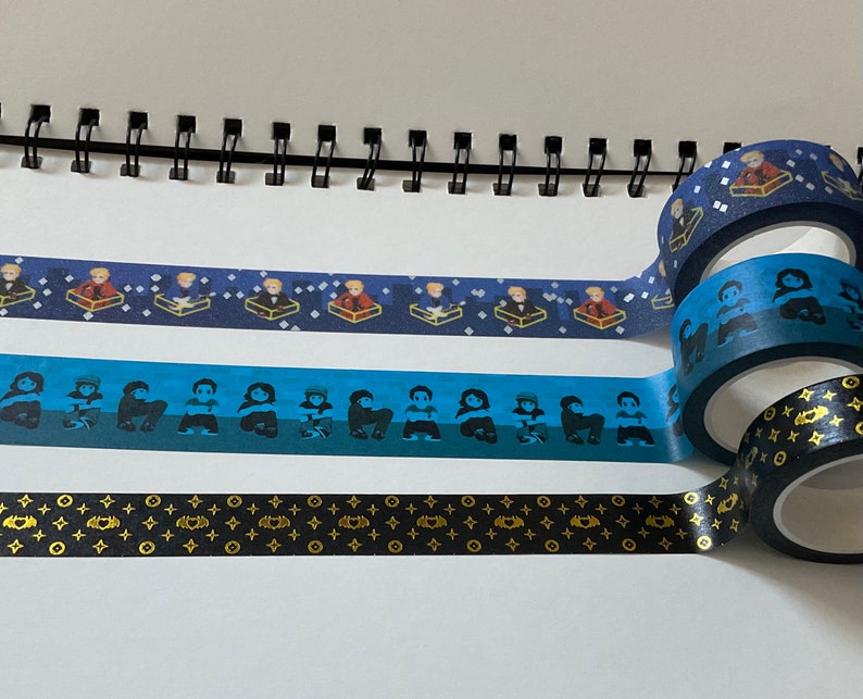 May include: Three rolls of blue and black patterned washi tape. The top roll has a pattern of small figures in boxes. The middle roll has a pattern of small figures. The bottom roll has a pattern of black and gold geometric shapes.