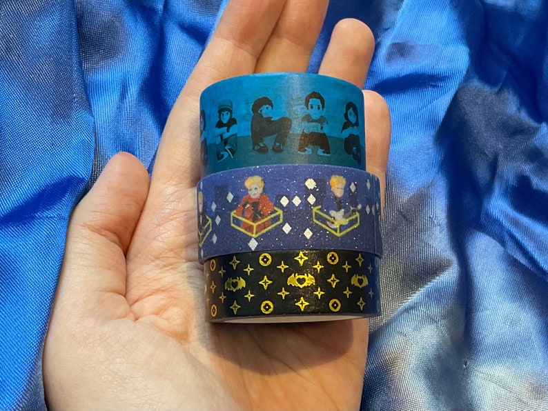 May include: Three rolls of washi tape featuring cartoon characters. The top roll has a blue background with white cartoon characters. The middle roll has a blue background with white and gold glitter and white cartoon characters. The bottom roll has a black background with gold stars and white cartoon characters.