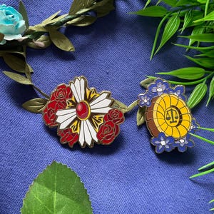 May include: Two enamel pins featuring floral designs. The first pin is a white flower with red roses and a red gem in the center. The second pin is a yellow circle with a scale of justice design surrounded by blue flowers.