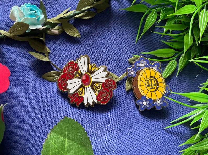 May include: Two enamel pins featuring floral designs. The first pin is a white flower with red roses and a red gem in the center. The second pin is a yellow circle with a scale of justice design surrounded by blue flowers.