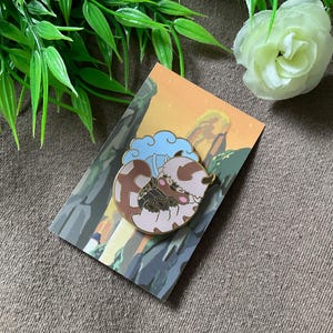 May include: A gold-coloured enamel pin featuring a cartoon character with brown fur, a white face, and blue horns. The character is curled up in a ball, with a blue cloud behind it. The pin is on a card with a background of a mountain range and a sunset.