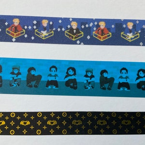 May include: Three different washi tapes with various designs. The first tape has a blue background with gold stars and a repeating pattern of a person sitting in a box. The second tape has a blue background with a repeating pattern of people sitting with their arms crossed. The third tape has a black background with a repeating pattern of gold stars and a bat with a heart in the center.