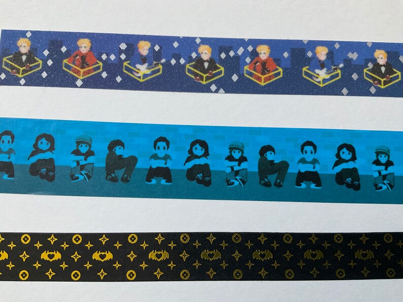 May include: Three different washi tapes with various designs. The first tape has a blue background with gold stars and a repeating pattern of a person sitting in a box. The second tape has a blue background with a repeating pattern of people sitting with their arms crossed. The third tape has a black background with a repeating pattern of gold stars and a bat with a heart in the center.