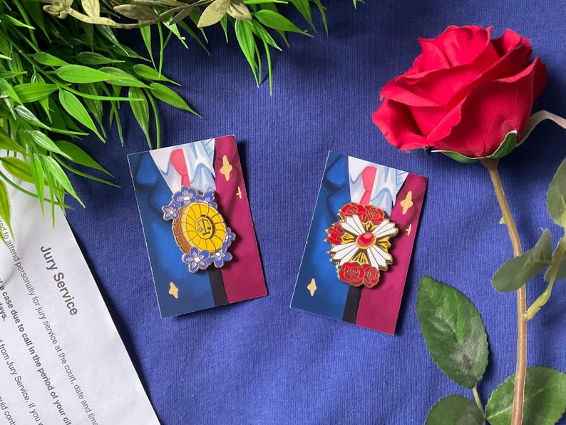 May include: Two enamel pins featuring a gold and blue design with a flower and a gold and red design with a flower. The pins are on a blue background with a red rose.