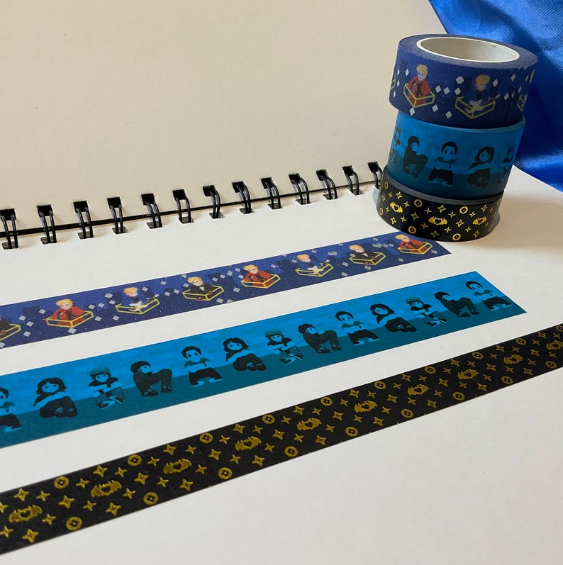May include: A stack of four washi tapes with different designs. The top tape is blue with a white and gold pattern. The second tape is blue with a white and black pattern. The third tape is blue with a white and gold pattern. The bottom tape is black with a gold pattern.