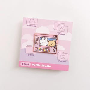Op de afbeelding: Een roze emaille pin met een witte konijn en een bruine beer in een roze computer venster. De pin zit op een roze kaart met de tekst "Start Puffie Studio". De kaart heeft illustraties van wolken en mappen.