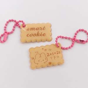 May include: Two keyrings with a cookie design and pink ball chains. One keyring reads "smart cookie" in a handwritten font. The other keyring has a cartoon drawing of a bear and the text "2+1=21 ???".