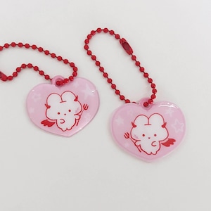 May include: Two pink heart-shaped keyrings with red ball chains. Each keyring features a white cartoon devil with red wings and a pitchfork. The background has white stars. The keyrings are on a white background.
