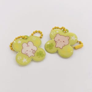 May include: Two clover-shaped keyrings with cartoon animal designs. One features a white bunny, the other a brown bear. Both are light green with gold-coloured ball chains. Each keyring has a clover, stars, and a cartoon animal.