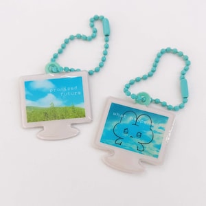 May include: Two computer-shaped keyrings with turquoise beaded chains. One keyring displays a landscape with the text "promised future." The other shows a cartoon face with the text "what happened." Both have a white frame.