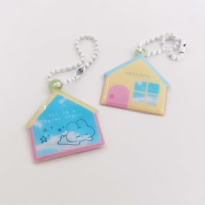 May include: Two house-shaped keyrings with white beaded chains. One keyring has a blue roof with the text "this is > fever dream" and a cloud illustration. The other has a yellow house with a pink door and the word "welcome".