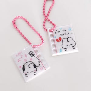 May include: Two clear acrylic keychains with pink ball chains. One keychain features a drawing of a music player and headphones, with musical notes and flowers. The other keychain has the text "I'm so tired..." and a broken heart, with a sad cloud face.