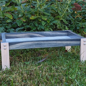 May include: A wooden tray with a blue and black stained top and natural wood legs. The tray has a white border around the top and is sitting on a green grassy surface.