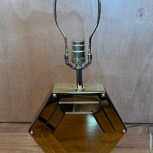 May include: A gold-toned table lamp with a geometric design. The lamp features a hexagonal base and a clear acrylic base. The lamp's metal frame supports a curved arm that holds the light bulb. The lamp is approximately 18 inches tall.