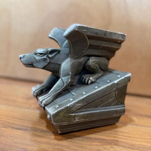 May include: A detailed, gray gargoyle statue with outstretched wings and a fierce expression. The statue is perched on a rectangular base with rivets, showcasing a weathered, antique finish. The sculpture is a decorative item.