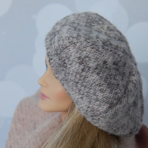Puede incluir: Una muñeca con un gorro de punto gris y una bufanda de punto rosa. La muñeca tiene el pelo largo y rubio.